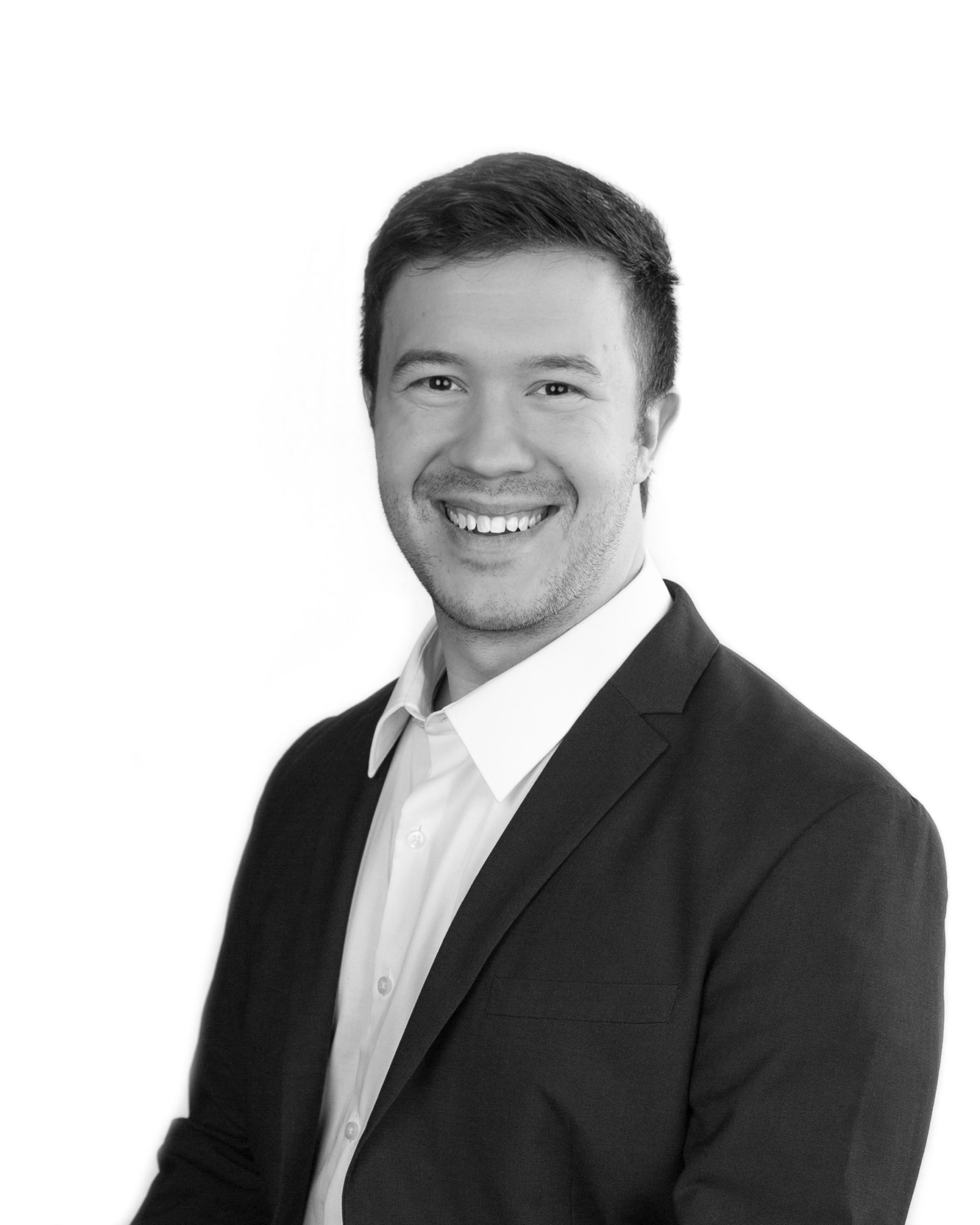 A man in a dark blazer and white shirt smiles against a plain white background. He has short, dark hair and a friendly expression. The image is in black and white.