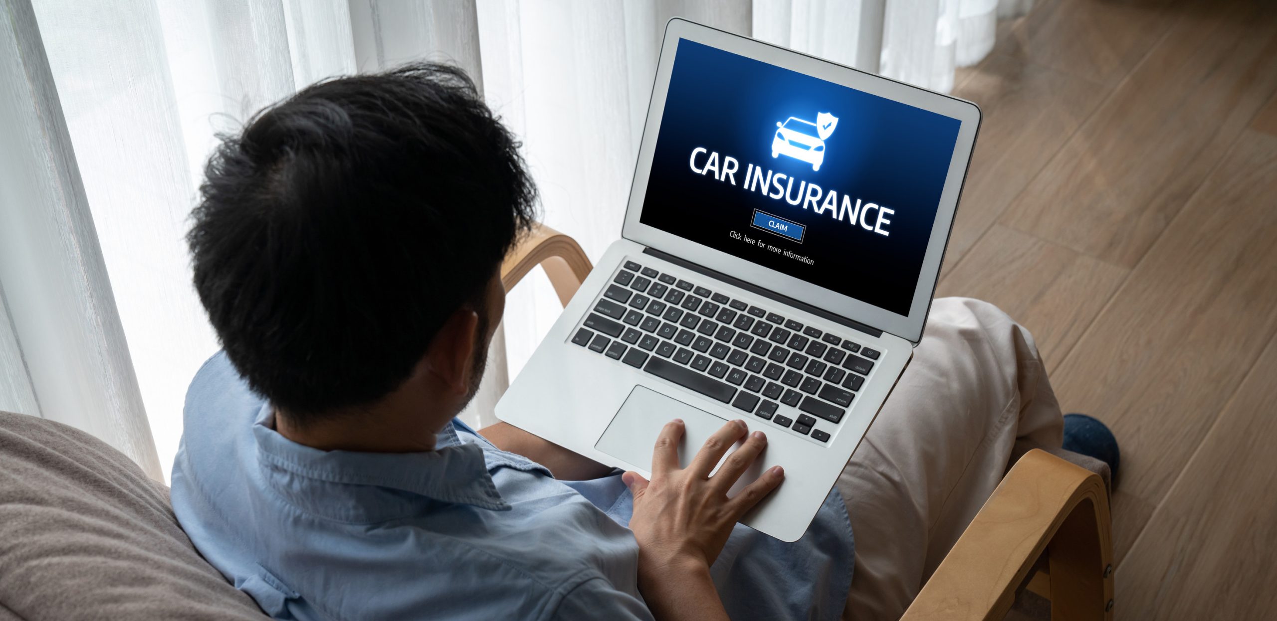 A person sitting on a chair uses a laptop displaying an “Auto Insurance 101” website with a car and shield icon, ready to click the “Start” button. Light from a window illuminates the scene.