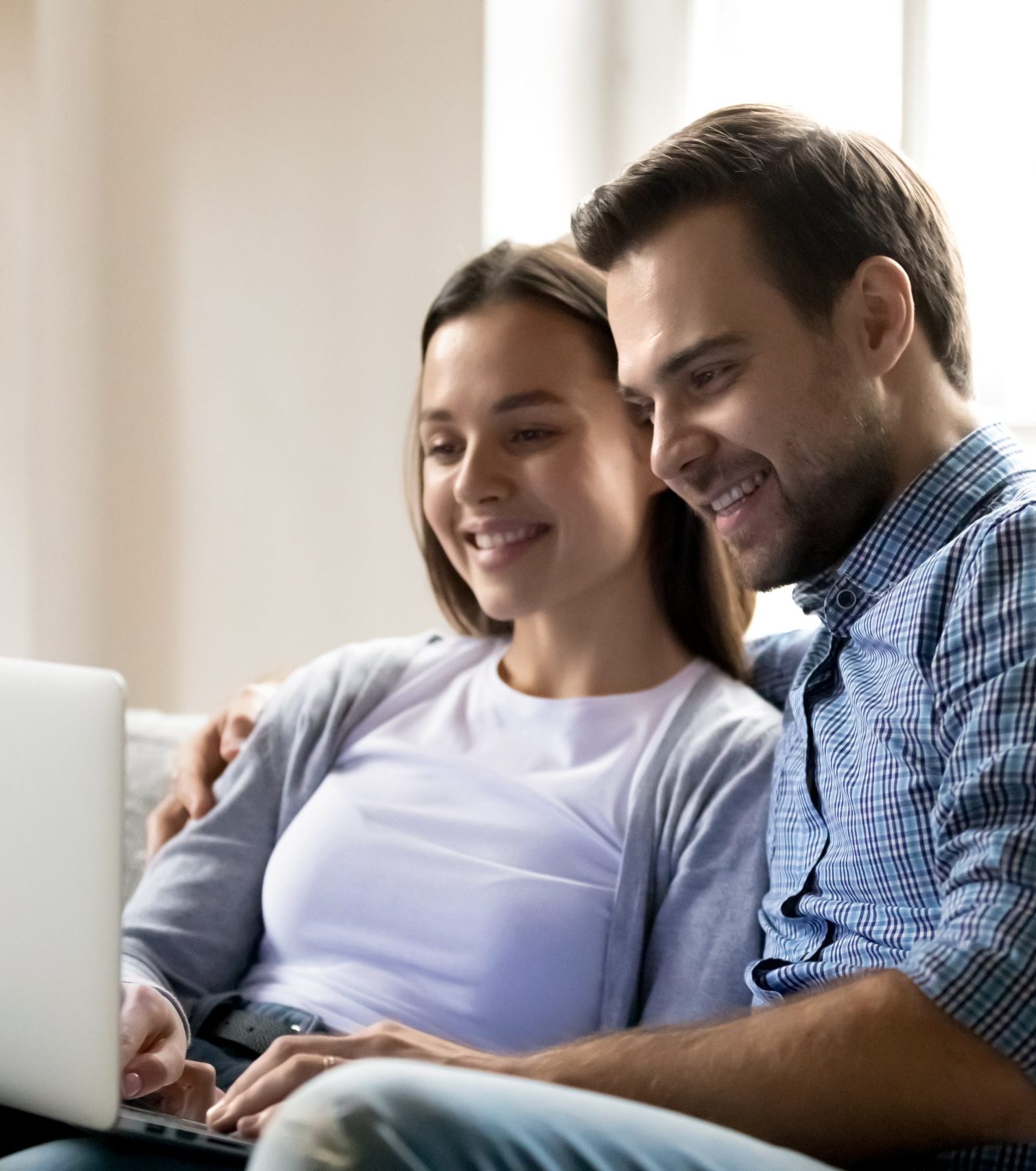 A smiling couple sits closely together on a couch, looking at a laptop screen. The woman is typing while the man has his arm around her, and they both seem happy and engaged with what they are viewing.