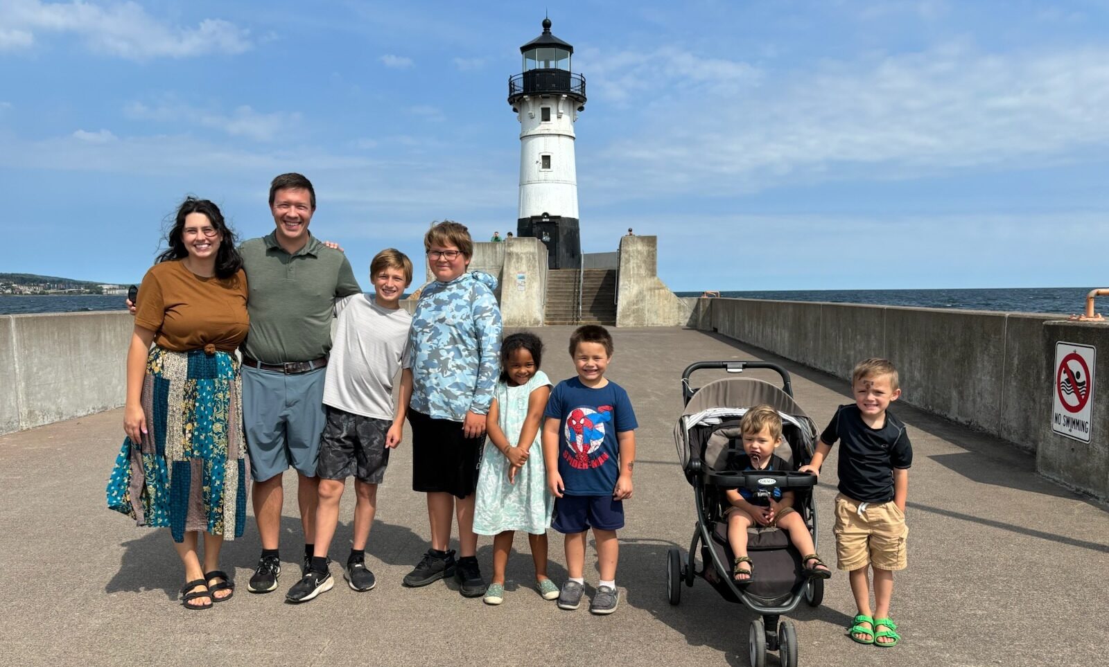A group of eight people, including adults and children, pose together on a pier in front of a lighthouse. One child sits in a stroller. The weather is sunny with a partly cloudy sky.