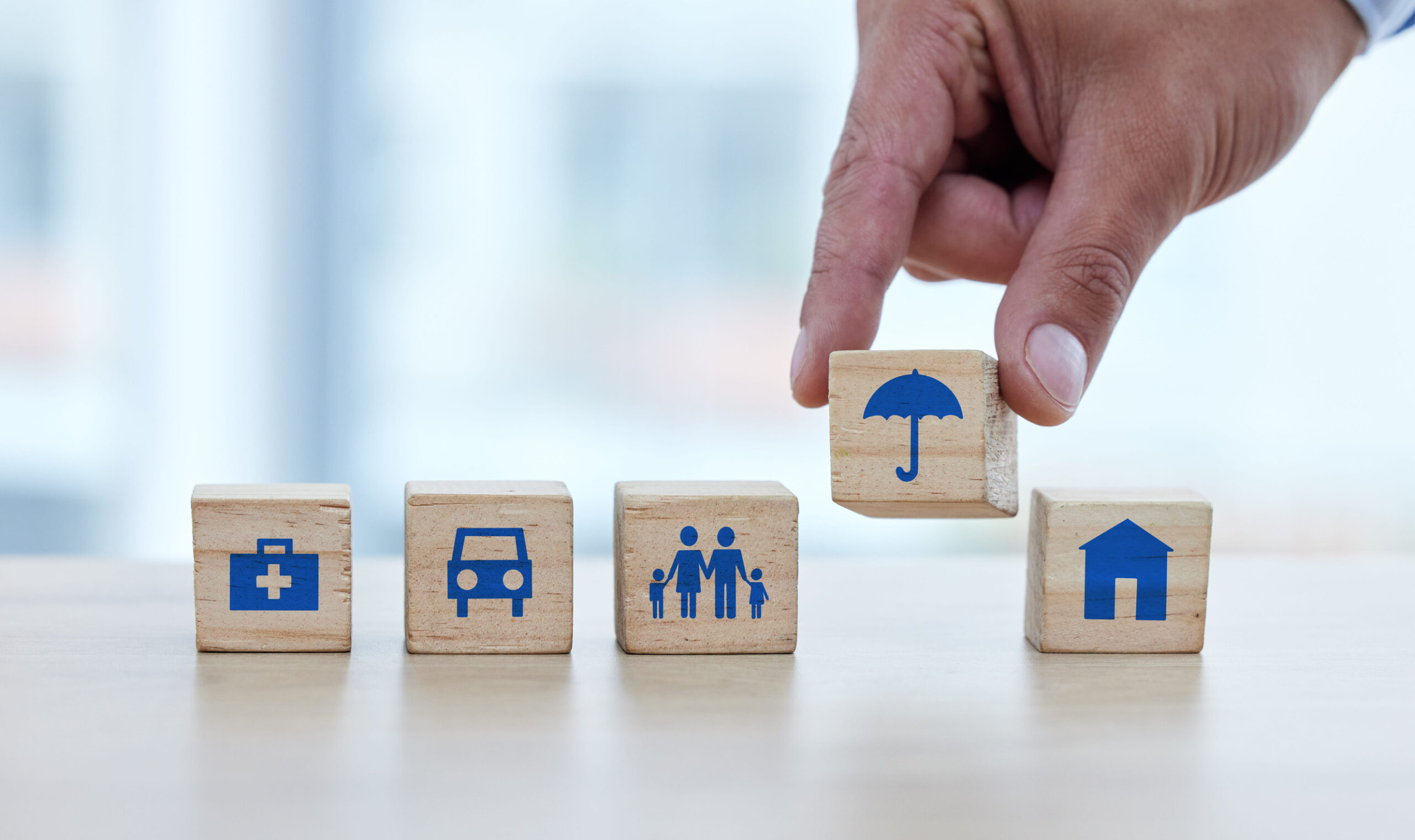 A hand arranges wooden blocks with blue icons representing health, car, family, umbrella (insurance), and home—visuals that highlight smart reasons to consult a reliable insurance broker for comprehensive coverage.