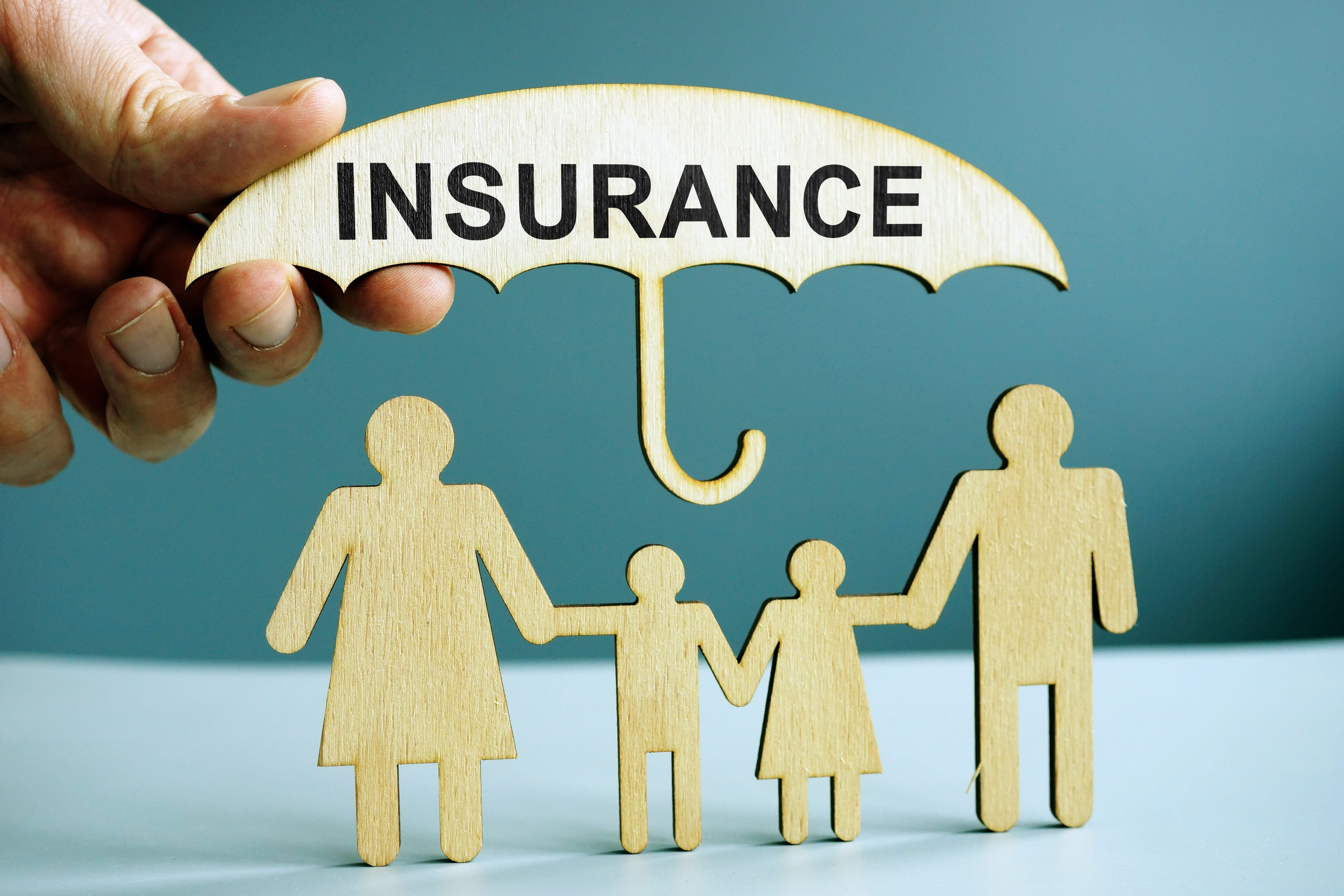 A hand holds a wooden umbrella labeled “INSURANCE COVERAGE” over wooden cutouts of a family with two adults and two children, symbolizing secure protection and the importance of safeguarding loved ones.