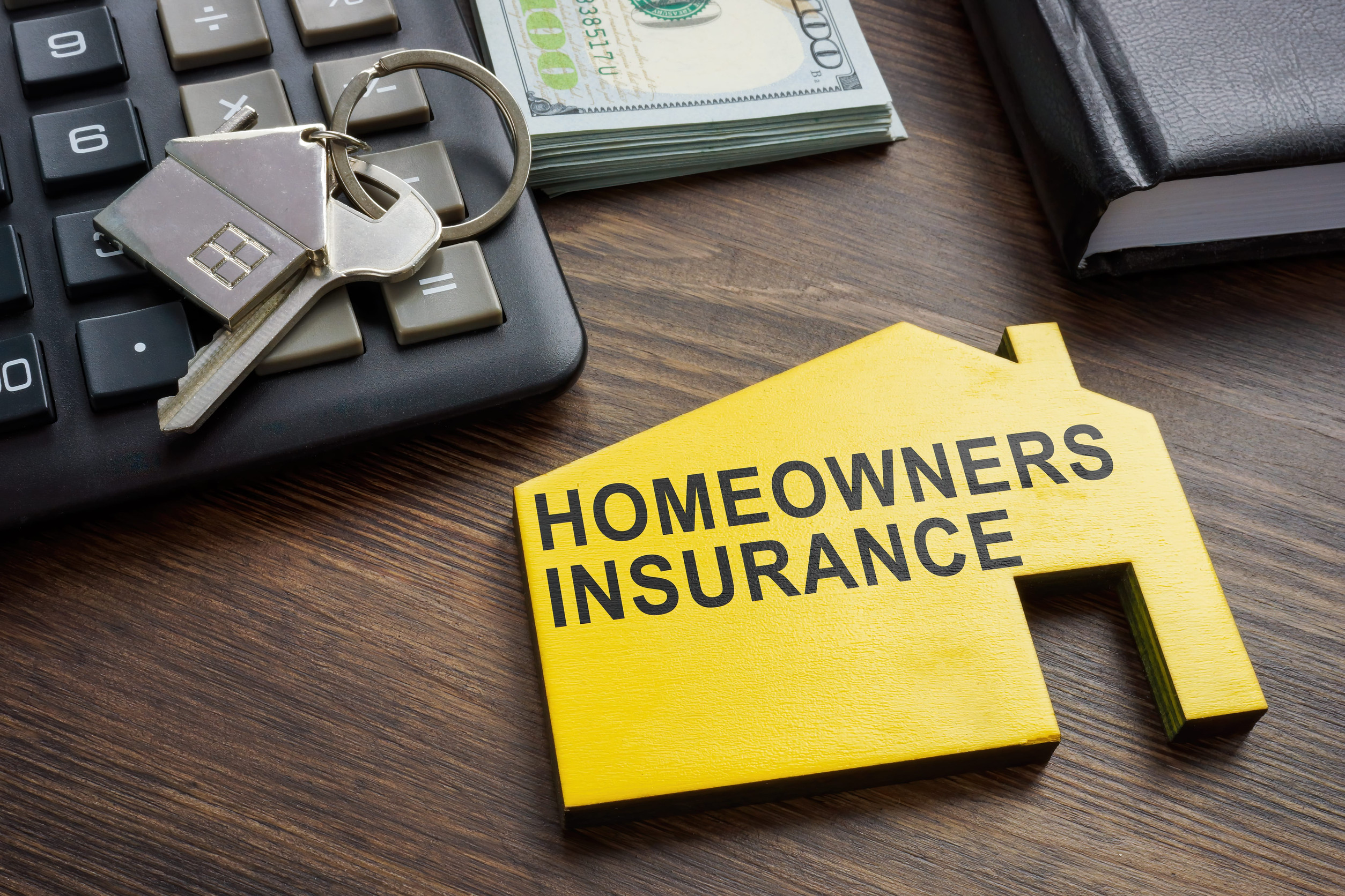 A yellow house-shaped sign with "Homeowners Insurance" text sits on a wooden desk beside house keys, a calculator, a stack of hundred-dollar bills, and a closed notebook—highlighting the importance of checking for Coverage Gaps.