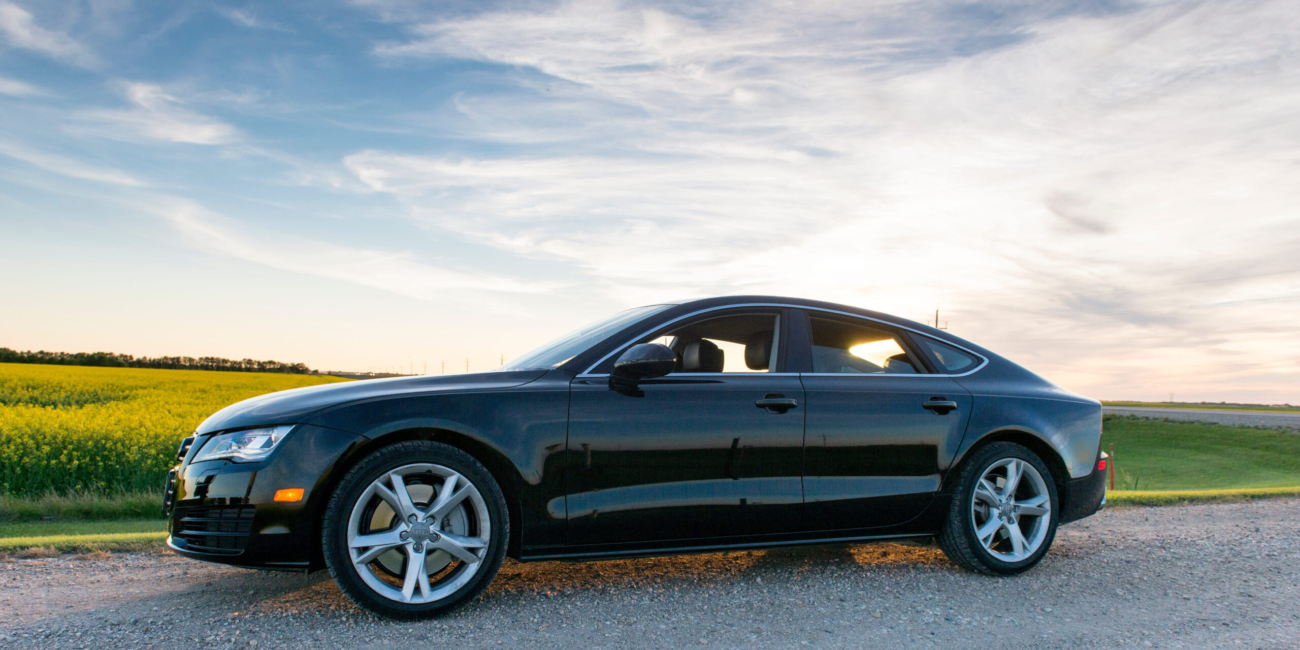 A sleek black car, covered on the road with auto insurance, is parked on a gravel road beside a green field under a blue sky with wispy clouds at sunset.