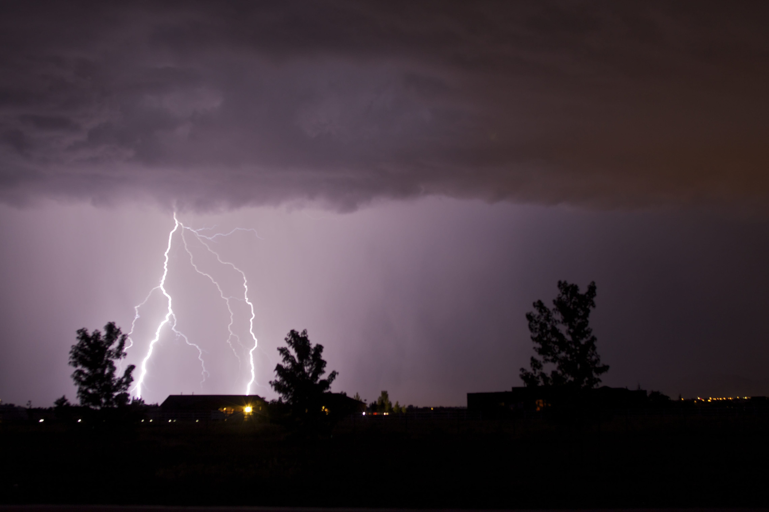 A dark, cloudy sky is lit up by multiple bright lightning bolts striking near silhouetted trees and buildings at night, highlighting the importance of storm damage insurance amid the scattered lights in the background.