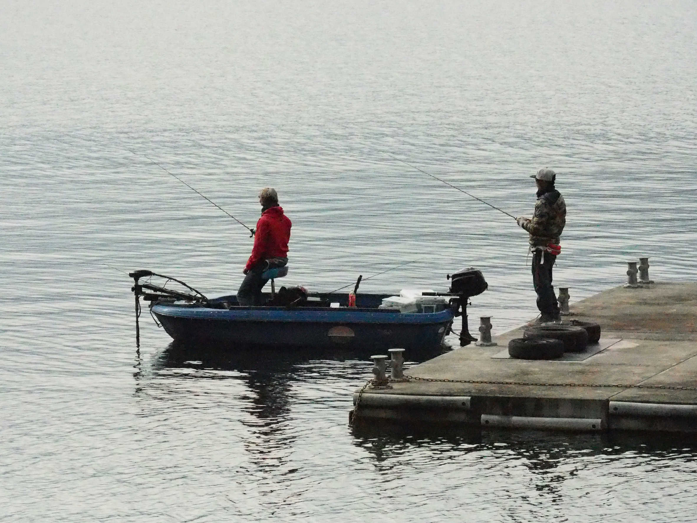Two people fishing: one stands on a small blue boat covered by watercraft liability insurance, the other on a dock beside the boat. Both are holding fishing rods over calm water, dressed warmly in jackets. The scene is peaceful and quiet.