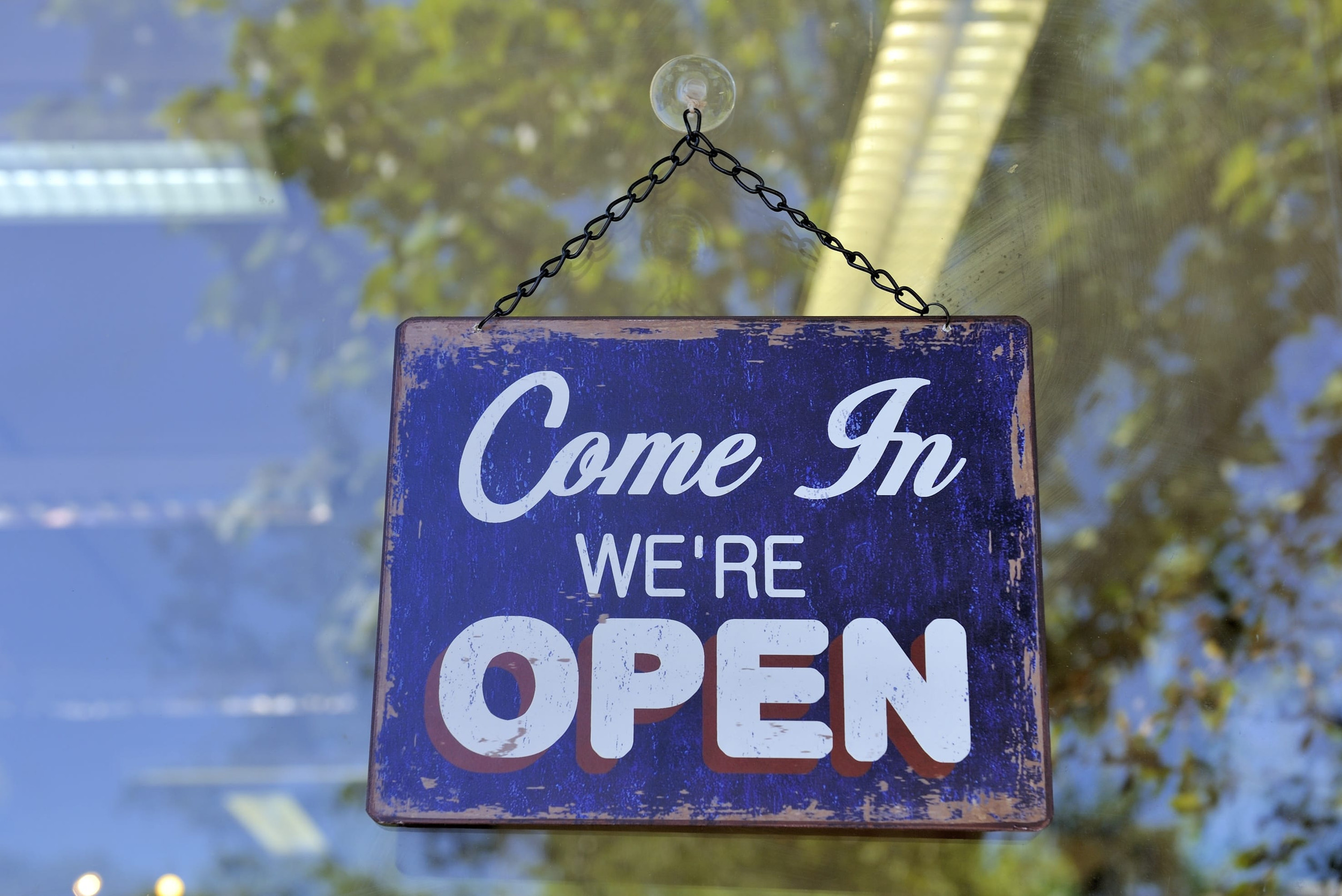 A blue and white sign hanging on a window reads “Come In WE’RE OPEN.” The background shows reflections of trees and ceiling lights—a welcoming scene that highlights the importance of commercial property insurance for your business.