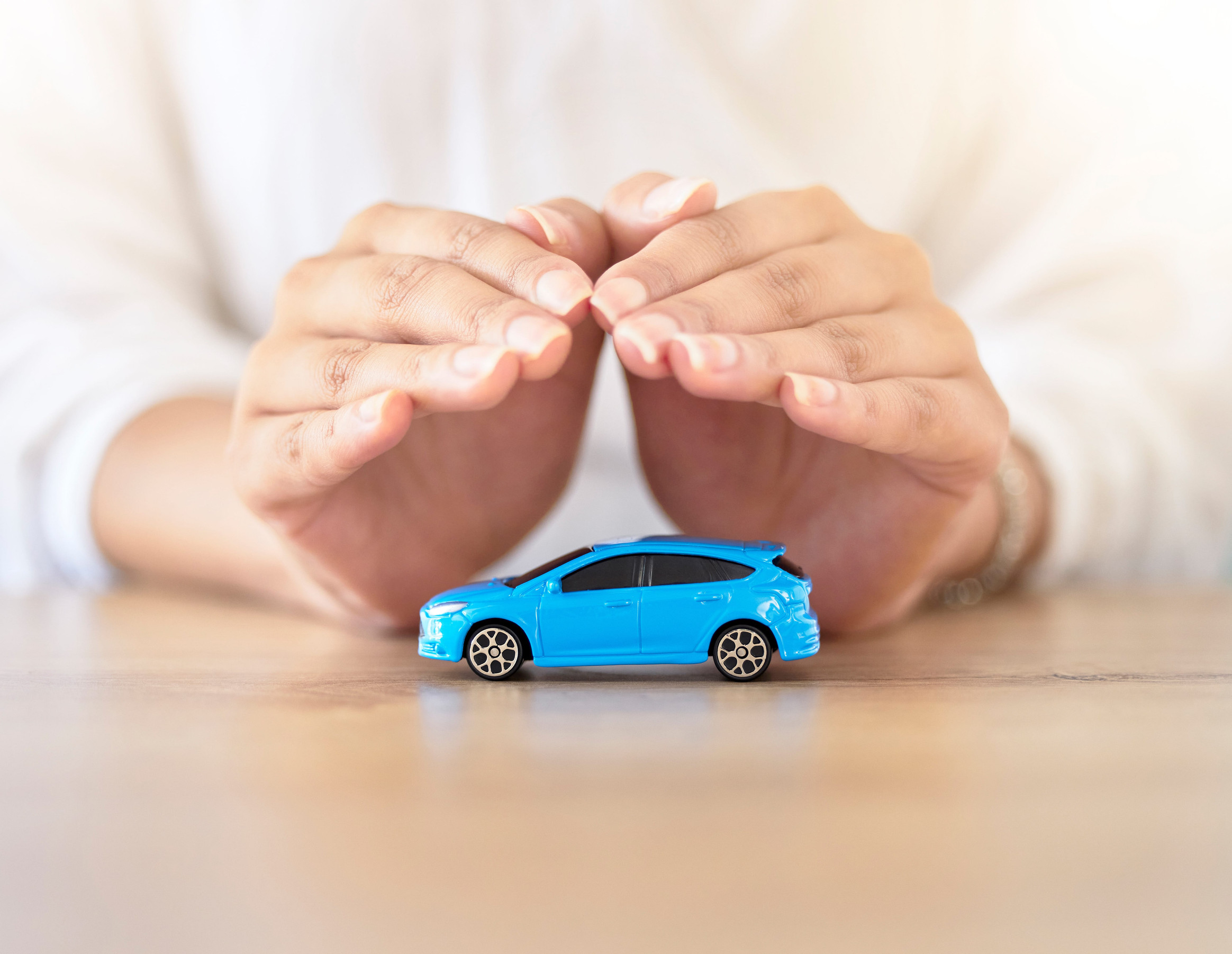 A person’s hands are cupped protectively over a small blue toy car on a wooden surface, symbolizing car insurance or vehicle protection—an image that resonates especially with new car owners seeking comprehensive insurance coverage.