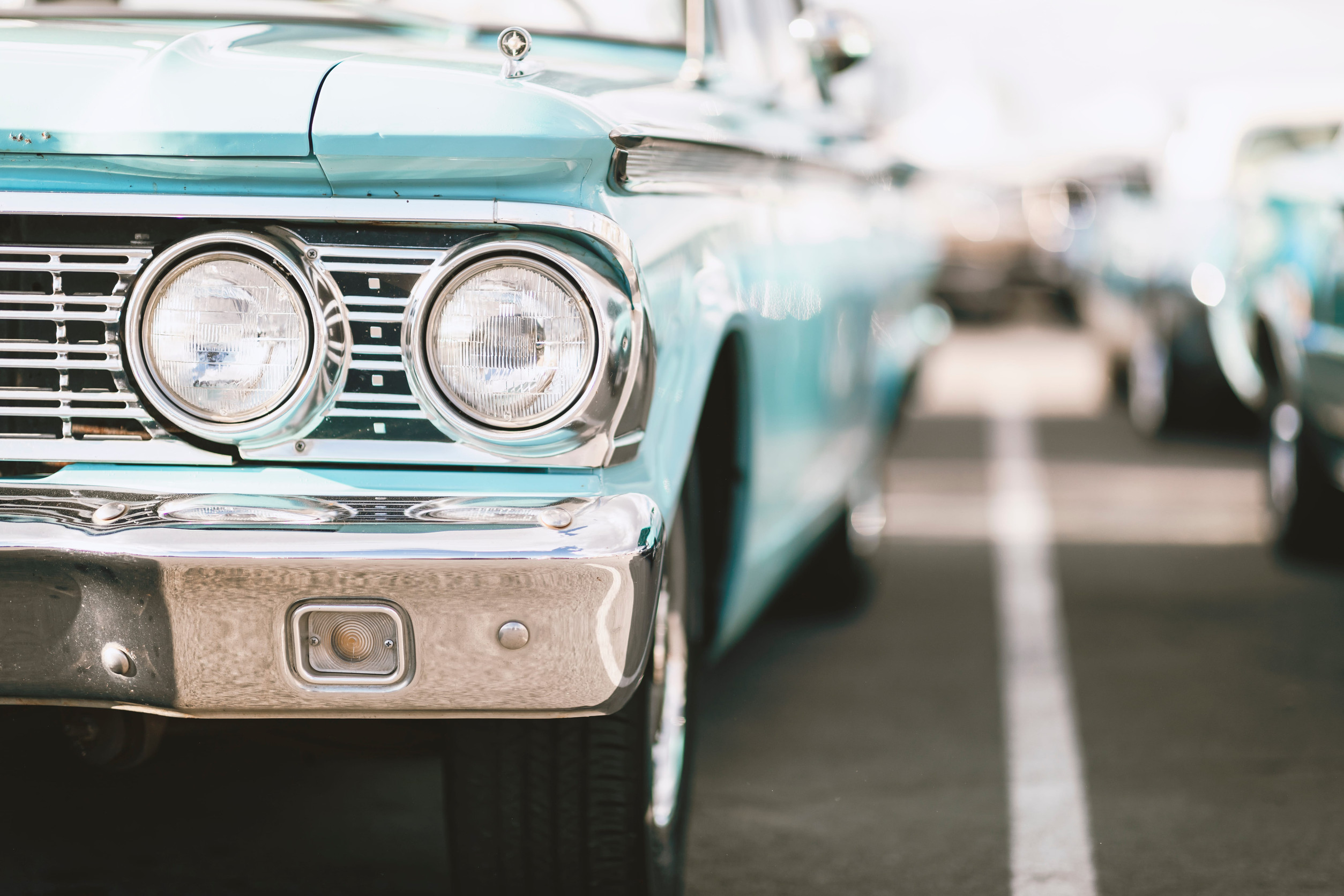 Close-up of the front left side of a classic car in vintage light blue, parked in a row, showing its headlights, grille, and chrome details under natural sunlight—a perfect snapshot for any classic car guide.